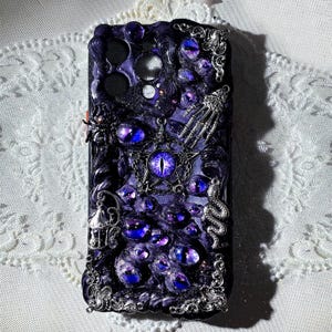 May include: A black phone case with a detailed design of purple eyes, a silver skeleton hand, and a silver snake. The case has a glittery finish and a silver pentagram with a large purple eye in the center.