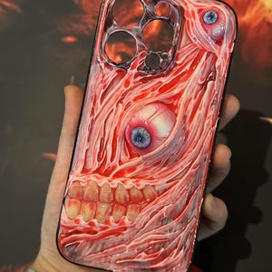 May include: A clear phone case featuring a red and white design that mimics exposed flesh, complete with teeth and eyeballs. The case has a black border and cutouts for the phone's camera and buttons. The design is detailed and textured.