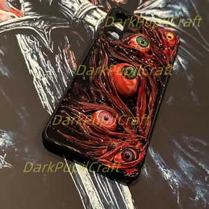 Puede incluir: Una funda de tel&eacute;fono negra con un dise&ntilde;o rojo y negro que presenta varios ojos. La funda tiene un aspecto texturizado, carnoso, con un fondo rojo oscuro y varios ojos rojos con pupilas negras.