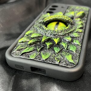 May include: A black smartphone case featuring a three-dimensional, textured design of green reptilian scales and a large, prominent central eye with a yellow iris and a vertical black pupil, set against a dark, matte-finished frame.