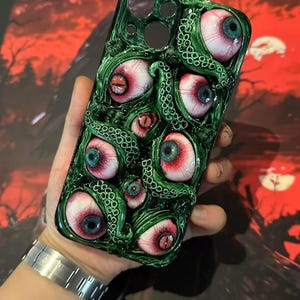 Op de afbeelding: Groene telefoonhoes met een gedetailleerd ontwerp van meerdere oogballen met rode en blauwe irissen en groene tentakels. De hoes heeft een glanzende afwerking en een unieke, artistieke uitstraling. De achtergrond is een wazig rood en zwart.