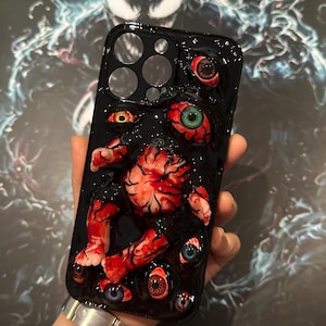 Pode incluir: Capa preta para iPhone com um gráfico de um bebê sangrento com vários olhos. A capa tem um fundo preto com detalhes vermelhos e brancos.