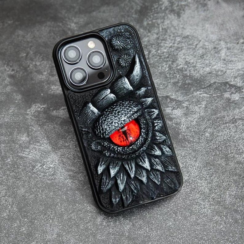 May include: A black smartphone case featuring a three-dimensional, textured dragon scale design. The center of the case displays a prominent, realistic dragon eye with a vibrant red iris and a vertical black pupil, surrounded by dark scales.