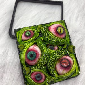 May include: A square compact mirror featuring a three-dimensional sculpted cover with a green, tentacled surface embedded with multiple realistic human-like eyes in various colors including green, blue, and red, set in a black frame.