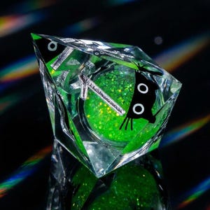 May include: A clear, faceted resin object with a green glitter interior. The object has a black butterfly design on one side and silver accents. The glitter is a vibrant green color, and the overall effect is eye-catching and unique.