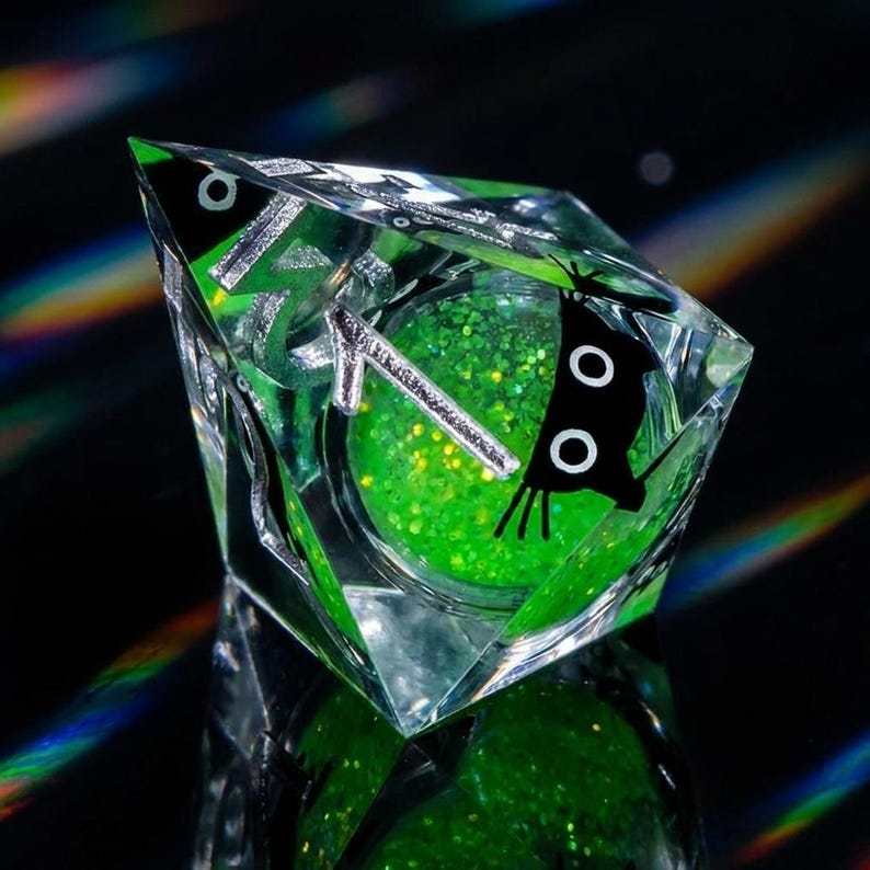 May include: A clear, faceted resin object with a green glitter interior. The object has a black butterfly design on one side and silver accents. The glitter is a vibrant green color, and the overall effect is eye-catching and unique.