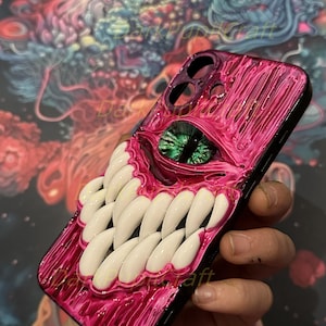 May include: A pink and black phone case with a 3D sculpted design of a monster with a large green eye and rows of white teeth.