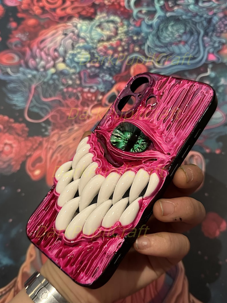 May include: A pink and black phone case with a 3D sculpted design of a monster with a large green eye and rows of white teeth.