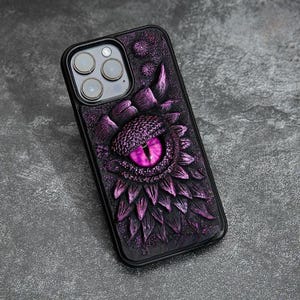 May include: A black smartphone case featuring a three-dimensional, textured design of a large, reptilian eye with a bright purple iris. The surface is covered in overlapping, scale-like purple and black plates surrounding the central eye.