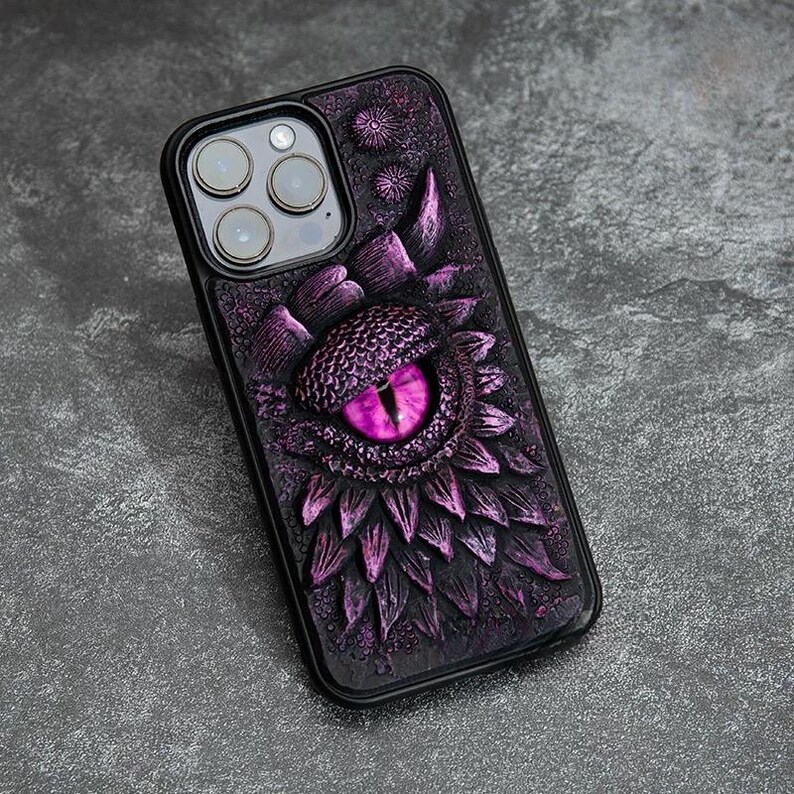 May include: A black smartphone case featuring a three-dimensional, textured design of a large, reptilian eye with a bright purple iris. The surface is covered in overlapping, scale-like purple and black plates surrounding the central eye.