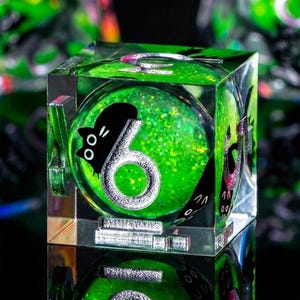 May include: A clear, square die with a green, glitter-filled interior. The number 6 is displayed in silver, with a black cat silhouette. The die is reflected on a mirrored surface, creating a unique visual effect.