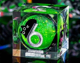 May include: A clear, square die with a green, glitter-filled interior. The number 6 is displayed in silver, with a black cat silhouette. The die is reflected on a mirrored surface, creating a unique visual effect.