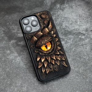 May include: A black smartphone case featuring a three-dimensional relief design of a dragon eye with a yellow iris and black pupil, surrounded by textured, bronze-colored scales and small floral accents on a dark, rugged background.