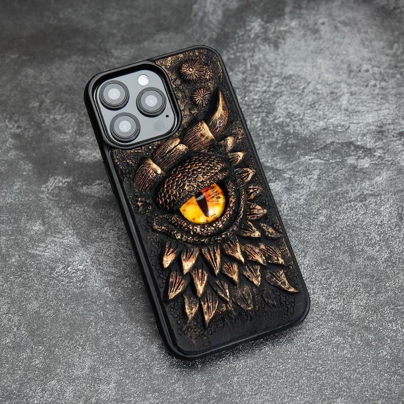 May include: A black smartphone case featuring a three-dimensional relief design of a dragon eye with a yellow iris and black pupil, surrounded by textured, bronze-colored scales and small floral accents on a dark, rugged background.