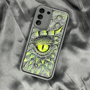 May include: A rectangular phone case featuring a detailed, textured graphic of a reptilian eye with a bright yellow iris and a vertical black pupil, surrounded by overlapping green and gray scales and spikes on a dark, textured background.