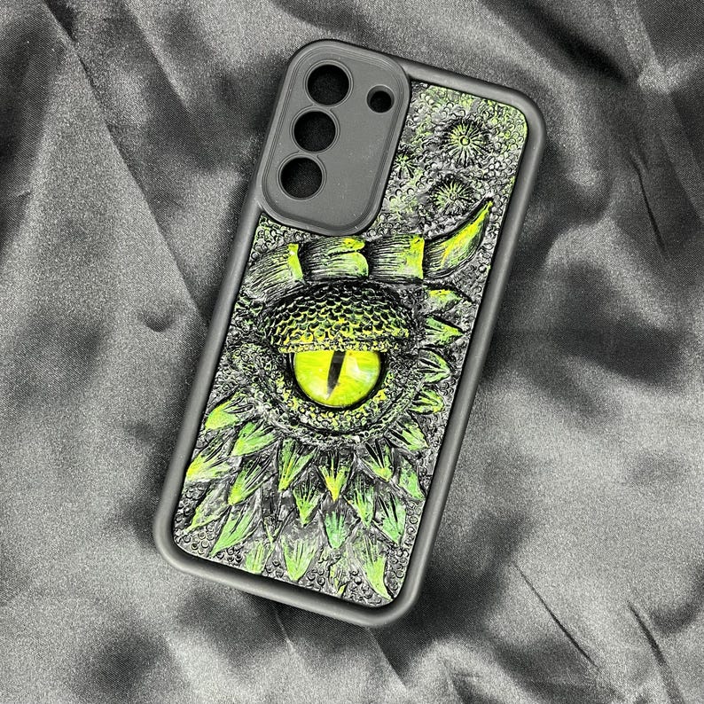 May include: A rectangular phone case featuring a detailed, textured graphic of a reptilian eye with a bright yellow iris and a vertical black pupil, surrounded by overlapping green and gray scales and spikes on a dark, textured background.