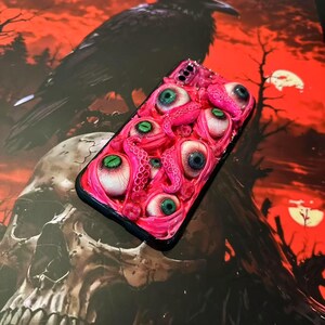 Op de afbeelding: Een roze telefoonhoesje met meerdere realistische oogballen en tentakels. De ogen hebben verschillende kleuren, waaronder groen en blauw. De hoes is geplaatst tegen een achtergrond van een raaf, schedel en een rode lucht.