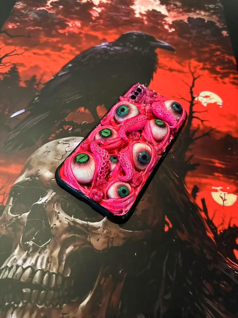 Op de afbeelding: Een roze telefoonhoesje met meerdere realistische oogballen en tentakels. De ogen hebben verschillende kleuren, waaronder groen en blauw. De hoes is geplaatst tegen een achtergrond van een raaf, schedel en een rode lucht.