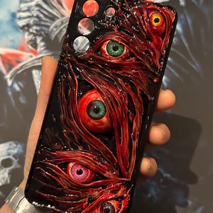 Puede incluir: Una funda de tel&eacute;fono negra con un dise&ntilde;o rojo, carnoso y texturizado. Cinco ojos con iris de diferentes colores est&aacute;n incrustados en la funda.