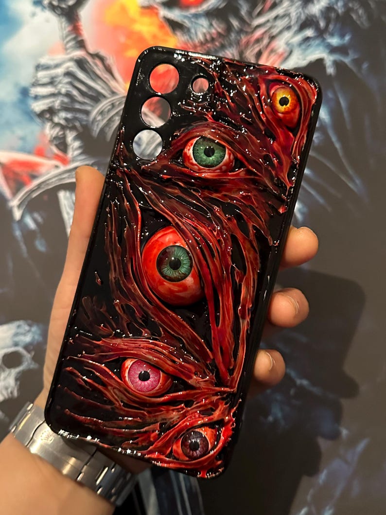 Puede incluir: Una funda de tel&eacute;fono negra con un dise&ntilde;o rojo, carnoso y texturizado. Cinco ojos con iris de diferentes colores est&aacute;n incrustados en la funda.