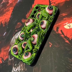 Op de afbeelding: Een groene telefoonhoes met meerdere realistisch ogende oogballen met rode en blauwe irissen, omringd door groene tentakels. De hoes heeft een glanzende afwerking en staat tegen een rode en zwarte achtergrond met een schedel en een raaf.
