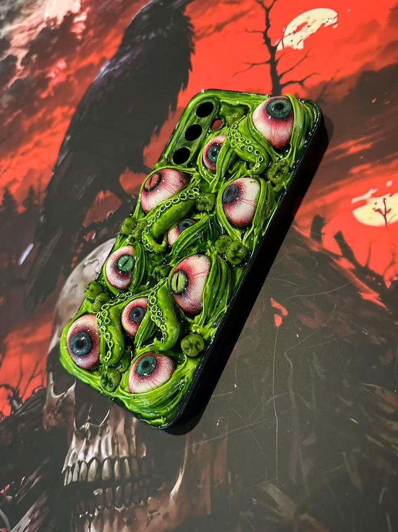 Op de afbeelding: Een groene telefoonhoes met meerdere realistisch ogende oogballen met rode en blauwe irissen, omringd door groene tentakels. De hoes heeft een glanzende afwerking en staat tegen een rode en zwarte achtergrond met een schedel en een raaf.