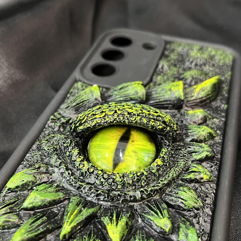 May include: A black smartphone case featuring a three-dimensional, textured design of a reptilian eye. The central eye has a bright yellow-green iris with a vertical black pupil, surrounded by raised, dark green and black scales.