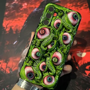 Op de afbeelding: Groene telefoonhoes met een gedetailleerd ontwerp van meerdere oogballen met rode, blauwe en groene irissen en groene tentakels. De hoes heeft een glanzende afwerking en een unieke, artistieke uitstraling.