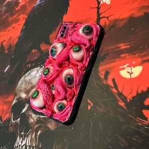 Op de afbeelding: Een roze telefoonhoesje met meerdere realistische ogen en tentakels. De hoes heeft een zwarte rand en een driedimensionaal ontwerp met textuur. De achtergrond is een donkere, rood-oranje sc&egrave;ne met een raaf en een schedel.