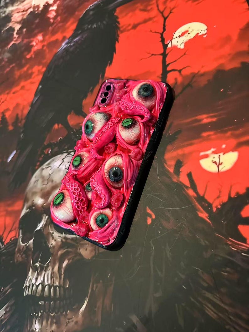 Op de afbeelding: Een roze telefoonhoesje met meerdere realistische ogen en tentakels. De hoes heeft een zwarte rand en een driedimensionaal ontwerp met textuur. De achtergrond is een donkere, rood-oranje sc&egrave;ne met een raaf en een schedel.