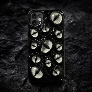 Könnte beinhalten: Eine schwarze Smartphone-Hülle mit einer texturierten, glänzenden Oberfläche, in die mehrere realistische, blassgrüne Augen mit dunklen vertikalen Pupillen eingebettet sind. Das Design bedeckt die gesamte Rückseite der Hülle.