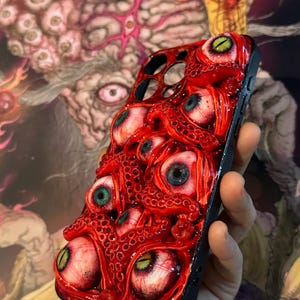 Op de afbeelding: Zwarte telefoonhoes met een gedetailleerd, driedimensionaal ontwerp met talrijke rode tentakels en oogballen met verschillende iris-kleuren. De hoes heeft een glanzende afwerking en een unieke, artistieke uitstraling.