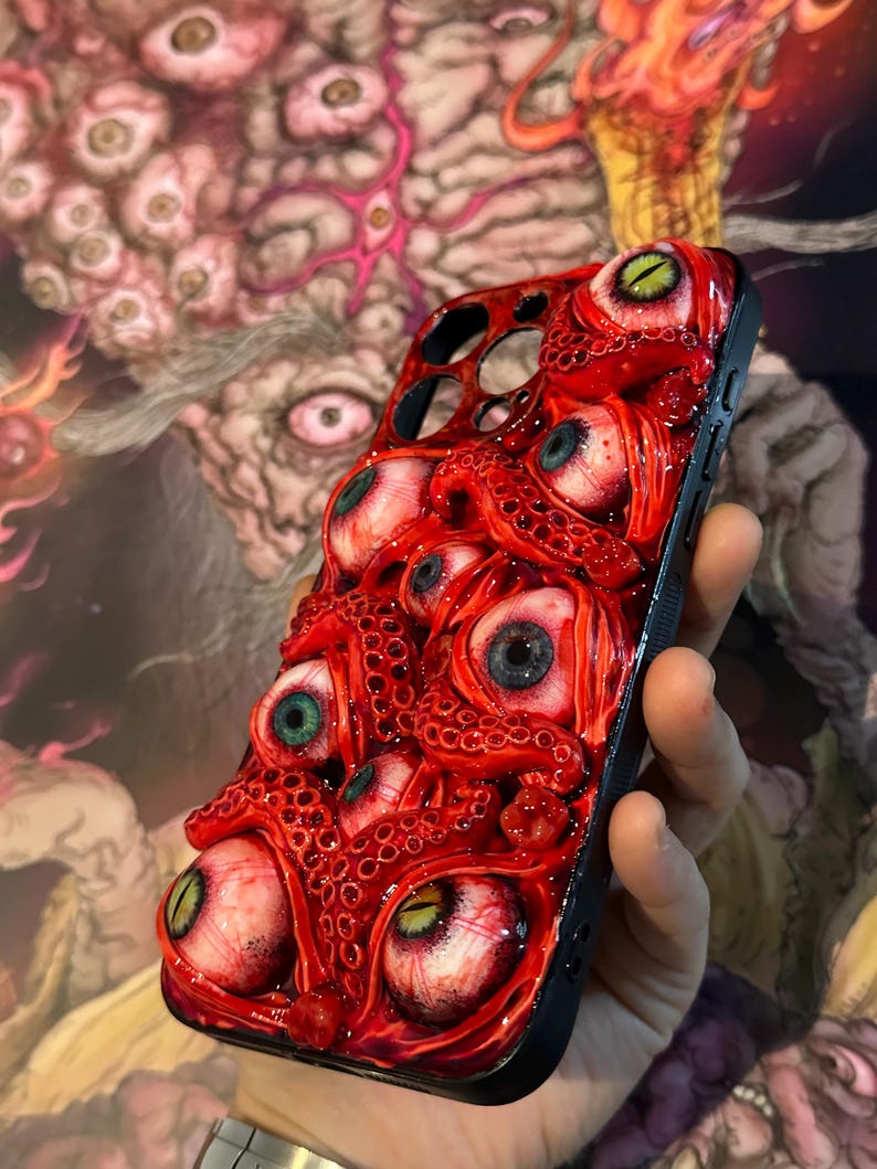 Op de afbeelding: Zwarte telefoonhoes met een gedetailleerd, driedimensionaal ontwerp met talrijke rode tentakels en oogballen met verschillende iris-kleuren. De hoes heeft een glanzende afwerking en een unieke, artistieke uitstraling.