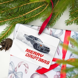 May include: A rectangular gift tag with a gray sports car illustration drifting on a snowy surface. The tag has the words "MERRY DRIFTMAS" in red. The tag is attached to a gift wrapped in patterned paper with a red ribbon.