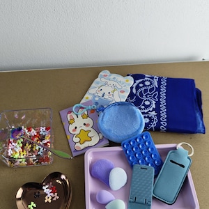 May include: A collection of small items including a blue bandana, a heart-shaped dish, a clear container with colorful confetti, and a pink tray with makeup sponges and a phone stand. There are also several small, decorative items with cartoon characters.