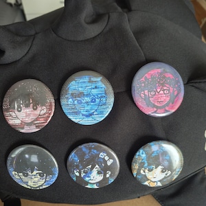 Op de afbeelding: Een zwarte, haai-vormige hoed met zes ronde, kleurrijke buttonspins. De buttons tonen illustraties van stripfiguren in rood, blauw en paars. De hoed is gemaakt van een zachte, getextureerde stof.