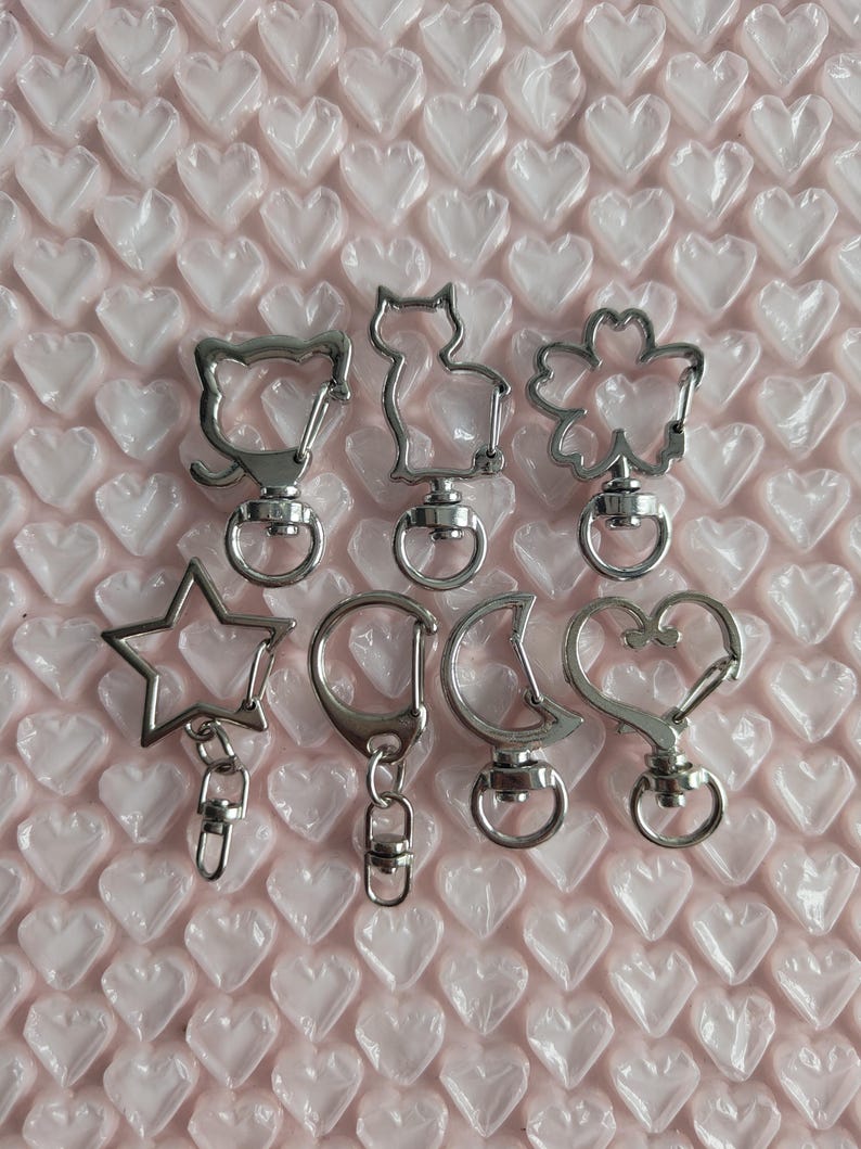 Op de afbeelding: Een verzameling zilverkleurige sleutelhangers in verschillende vormen: een kat, een bloem, een ster, een cirkel, een halve maan en een hart. De sleutelhangers liggen op een roze ondergrond met een hartpatroon.