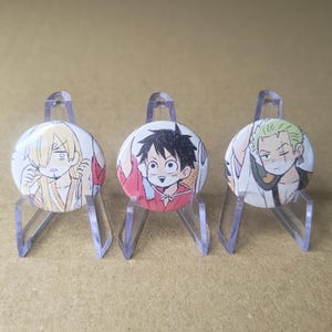 May include: Three circular pin-back buttons featuring anime-style character illustrations, each displayed on a clear plastic easel stand. The buttons depict characters with blonde hair, black hair, and green hair, respectively, in a colourful graphic art style.
