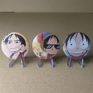 Puede incluir: Tres chapas redondas con ilustraciones de un personaje de estilo anime. La chapa de la izquierda muestra a un personaje con sombrero de paja sonriendo. La central muestra al personaje con gafas de sol pixeladas. La de la derecha tiene expresión de sorpresa.