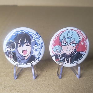 Könnte beinhalten: Zwei runde Ansteckbuttons mit Anime-Charakterillustrationen. Der linke Button zeigt eine Figur mit dunklem Haar in einem blauen Kapuzenpullover mit Sternenmotiven. Der rechte Button zeigt eine Figur mit hellblauem Haar und roten Kopfhörern.