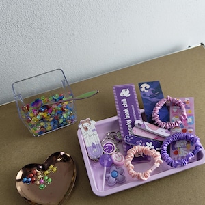 May include: A collection of accessories including a heart-shaped dish, a clear container with a spoon, and a purple tray. The tray holds hair ties, keychains, and stickers. The text "spooky book club" is visible on a purple label.