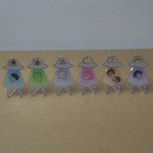 Op de afbeelding: Zes acryl bedels in de vorm van vliegende schotels, elk met een unieke karakterillustratie aan de binnenkant. De bedels hebben gekleurde doorschijnende lichamen in blauw, groen, paars, roze, geel en lavendel en staan op kleine doorzichtige standaards.