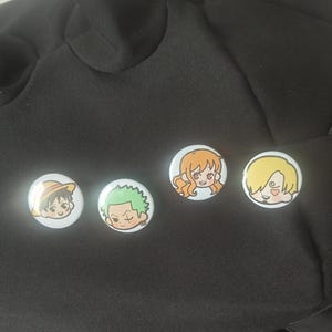 Könnte beinhalten: Vier runde Ansteckbuttons mit farbenfrohen Illustrationen von Anime-Charakteren im Chibi-Stil. Die Buttons zeigen eine Figur mit Strohhut, eine Figur mit grünen Haaren, eine mit orangefarbenen Haaren und eine blonde Figur mit einem Herz.