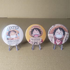 Könnte beinhalten: Drei runde Ansteckbuttons mit Monkey D. Luffy aus One Piece. Der linke Button ist weiß mit einem Steckbrief-Design. Der mittlere Button ist gelb und zeigt Luffy mit einem Wanted-Schild. Der rechte Button zeigt ein Porträt von Luffy.