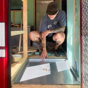 May include: A person is crouching down and inspecting a metal shower pan that is being installed in a small bathroom. The shower pan is rectangular and has a drain in the center. The bathroom is unfinished and has exposed wood framing.