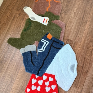 May include: A collection of handmade rugs in the shape of clothing items. Includes a brown pair of pants, a green jacket, blue jeans, a white t-shirt, red heart-patterned shorts, and a white sock.