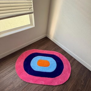 May include: A pink, oval-shaped rug with concentric circles in navy blue, light blue, and orange. The rug is placed on a dark wood floor near a window with blinds.