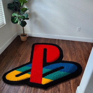May include: A multi-coloured rug in the shape of the PlayStation logo. The rug has a red 'P' shape, with blue, green, and yellow sections, all outlined in black. It's placed on a wooden floor.