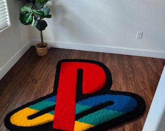 May include: A colorful rug in the shape of the PlayStation logo. The rug features a red 'P' shape, with blue, green, and yellow sections, all outlined in black. It's placed on a wooden floor.