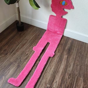 Pode incluir: Um tapete rosa vibrante em forma de uma figura caprichosa com cabeça, tronco e pernas longas. A cabeça apresenta um olho azul e uma expressão sorridente. O tapete é colocado sobre um chão de madeira, adicionando um toque lúdico à decoração.